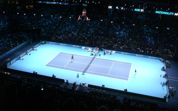 Ein Tennisplatz mit zwei Spielern, Bannern an den Seiten, einer sitzenden Menge im Stadion, einem Bild einer Person oben und Video-Kameras.