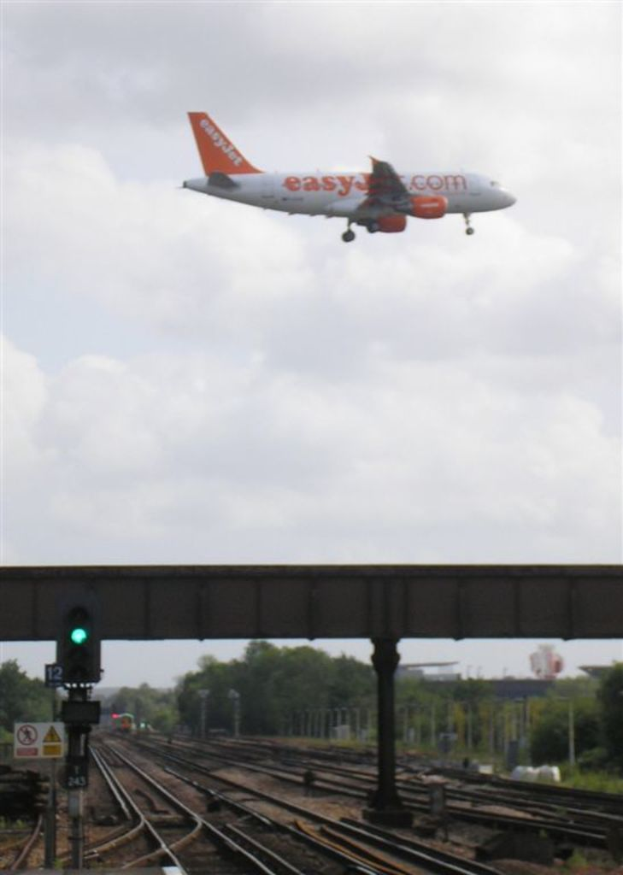 Eine Eisenbahnschiene mit einem Signalmast, einem Schild, einer Überführung und Bäumen im Vordergrund, mit einem Flugzeug am Himmel darüber.