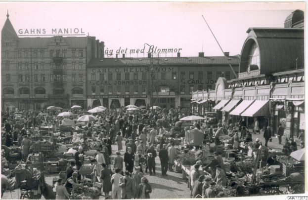 Schwarzes und weißes Foto eines belebten Berliner Markts mit Menschen, Gemüsestöllen und Gebäuden im Hintergrund.