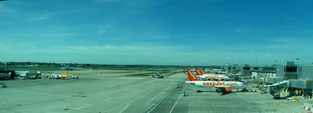 Eine Luftaufnahme eines Flughafens mit Flugzeugen, Gebäuden, Polen, Fahrzeugen, Bäumen und einer Straße unten sowie einem Zaun im Vordergrund.