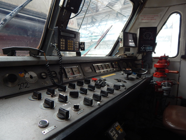 Eine Zugsteuerung mit Knöpfen, Schaltern und Messgeräten im Vordergrund und Fenstern an den Seiten, mit einem Zug im Hintergrund.