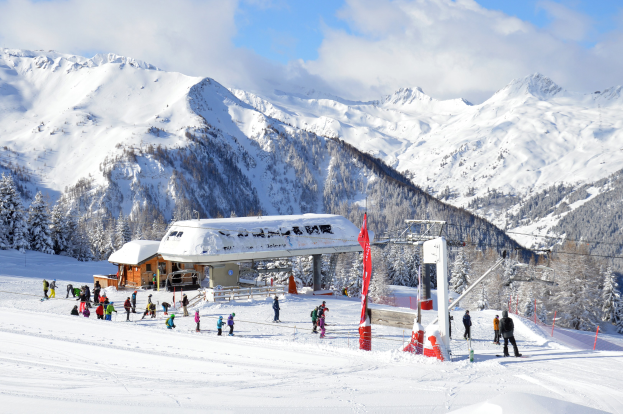 Gruppe von Menschen, die eine schneebedeckte Piste im österreichischen Alpengebiet mit Bäumen, Pfählen, Flaggen und einer Hütte im Hintergrund hinunterskifahren, unter einem bewölkten Himmel mit sichtbaren Bergen.