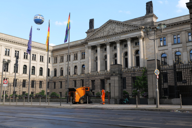 Großes modernes Gebäude mit zahlreichen Fenstern, als Bundestag in Berlin, Deutschland identifiziert, umgeben von Straßeninfrastruktur, Fußgängern und Flaggen, mit einem bewölkten Himmel im Hintergrund.