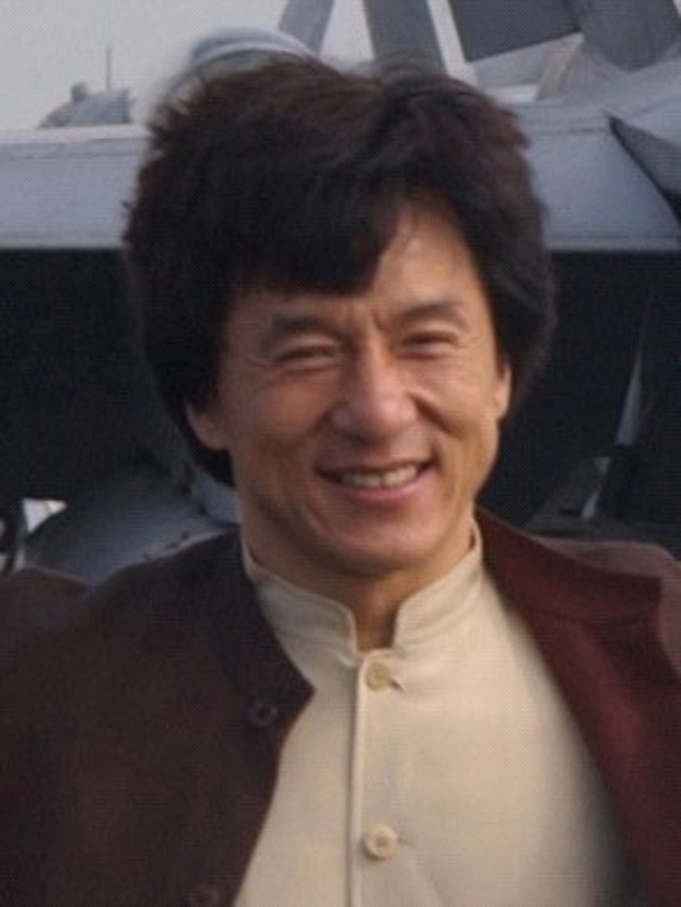 Jackie Chan steht lachend vor einem Flugzeug gegen einen klaren blauen Himmel, trägt ein weißes Hemd, eine schwarze Jacke und hat ordentlich gekämmtes Haar.