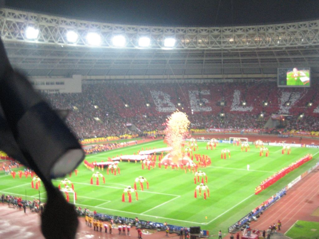 Ein Stadion mit Flutlicht, Eisengittern, Zuschauern, stehenden Personen, Werbetafeln, Anzeigetafeln, Sicherheitskameras, Flaggen und Dekorationen.