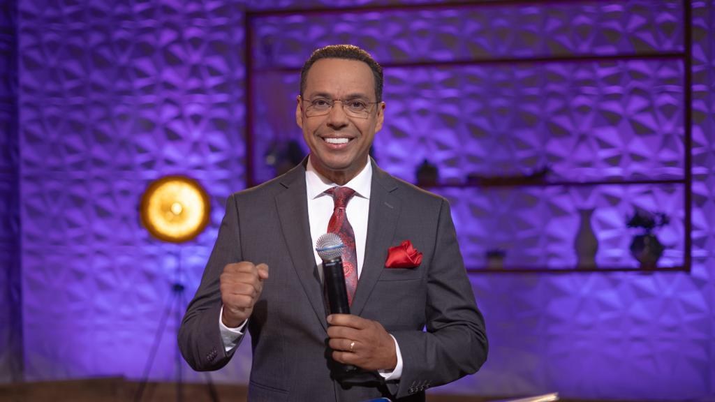 Stephen Colbert, Moderator von *The Daily Show with Trevor Noah*, steht auf einer Bühne und hält ein Mikrofon, trägt einen Anzug, Krawatte und eine Brille, mit einem Scheinwerfer und Ständer hinter ihm.