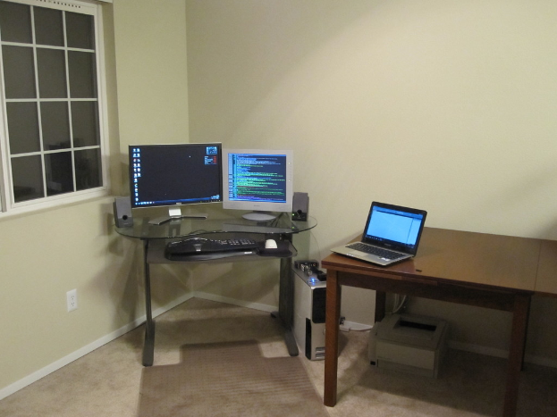 Ein Laptop auf einem Tisch mit einem darunter befindlichen Drucker, umgeben von Monitoren, einer Tastatur, einem mobilen Gerät und einer CPU, mit einem Fenster im Hintergrund.