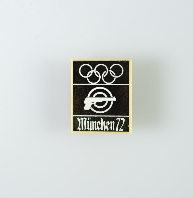 Ein schwarzes und weißes Logo mit einem schwarzen Kreis mit weißer Umrandung, der eine weiße Silhouette einer Person mit ausgestreckten Armen enthält, sowie der Text "München 1972" in fetter schwarzer Schrift.