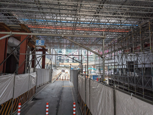 Baustelle mit ausgedehnten Gerüsten, Straße, Zaun, Infotafel, Metallrahmen, benachbarte Gebäude mit Fenstern und bewölktem Himmel.