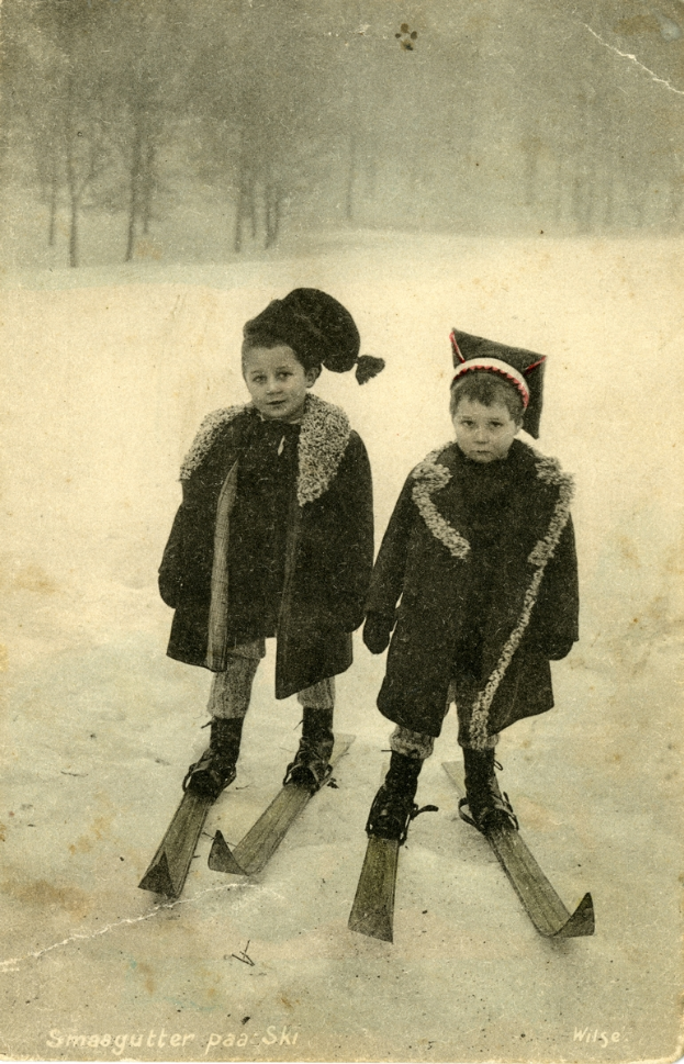 Zwei Kinder auf Skiern im Schnee, umgeben von Bäumen im Hintergrund, stehen auf dem schneebedeckten Boden.