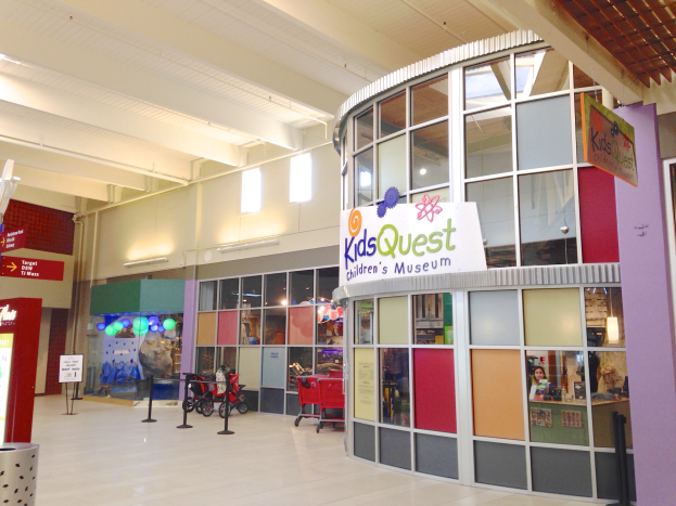 Innenraum eines Einkaufszentrums mit einem "Kids Quest Children's Museum"-Schild, Trolleys, ribbonierten Polen, Texttafeln, Deckenleuchten und verstreuten Gegenständen.