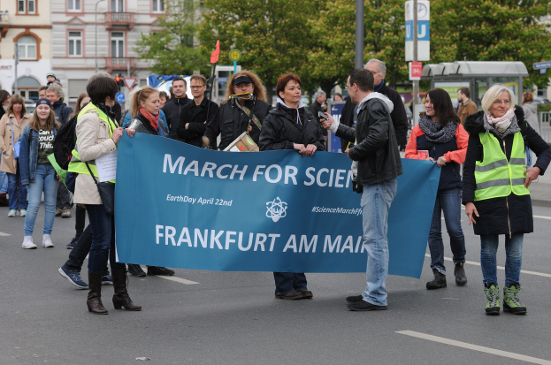 Gruppe von Menschen marschiert mit einem "March for Science Frankfurt am Main"-Schild die Straße entlang, Bäume und Gebäude im Hintergrund bei klarem blauem Himmel.