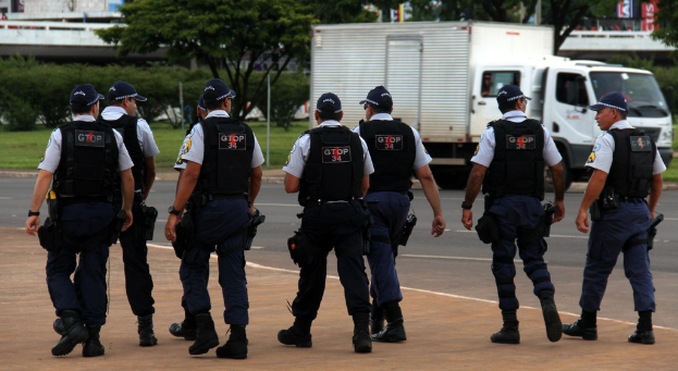 Eine Gruppe von Beamten in Uniform, einschließlich Mützen, Hemden, Schutzwesten, Uhren, Hosen und Schuhen, geht auf einer Straße. Rechts daneben steht ein Lastwagen in der Nähe eines Zauns, mit Gebäuden, Bäumen, Pflanzen, Polen und Bannern im Hintergrund und einer Brücke oben rechts.