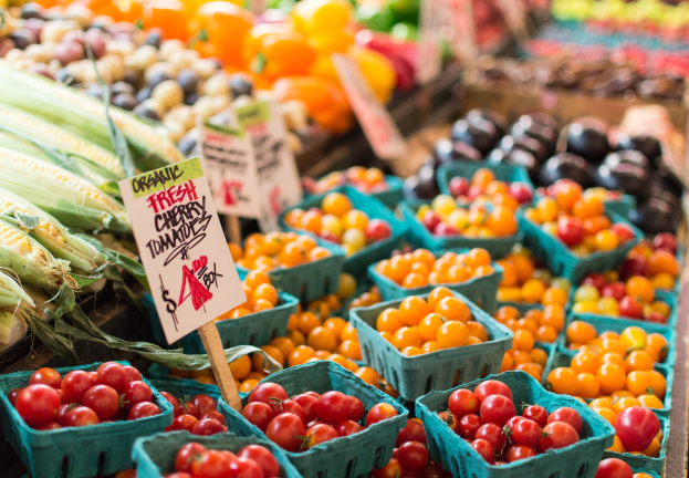 Frisches Obst und Gemüse, einschließlich Tomaten und Mais, in Körben auf einem Bauernmarkt mit Texttafeln im Hintergrund.