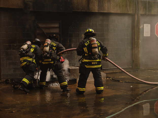 Feuerwehrleute in Helmen und Schutzausrüstung nutzen einen Schlauch, um ein Feuer zu löschen, während sie in der Nähe von Rohren, einer Wand, einer Tür und einem Schild stehen, mit Rauch im Hintergrund.