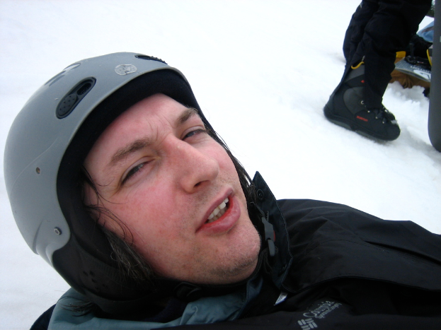Ein Mann in einem Pullover und Helm steht im Schnee, wobei die Beine und Skier einer anderen Person in der oberen rechten Ecke sichtbar sind.