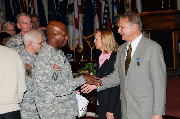 Eine Gruppe von Menschen, die mit zwei Personen Handschlag machen, Flaggen im Hintergrund auf Stangen, ein Podium mit einem Meilenstein und eine Topfpflanze in der Nähe.