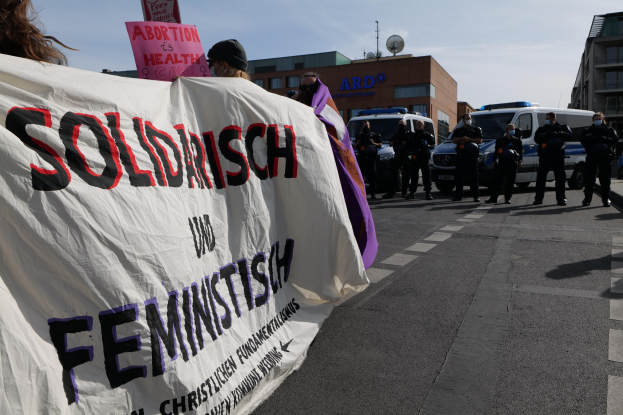 Gruppe von Menschen marschiert die Straße entlang und hält ein 'Solidarität und Feminismus'-Schild, mit geparkten Fahrzeugen, Gebäuden, einer Schüsselantenne und einem klaren blauen Himmel im Hintergrund.