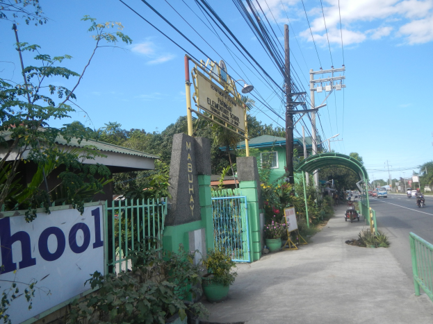 Schuleingangsschild am Straßenrand mit einem Bogen, Tor und Namensschild, umgeben von Fahrzeugen, Pflanzen, Bäumen, Strommasten und einem bewölkten Himmel.