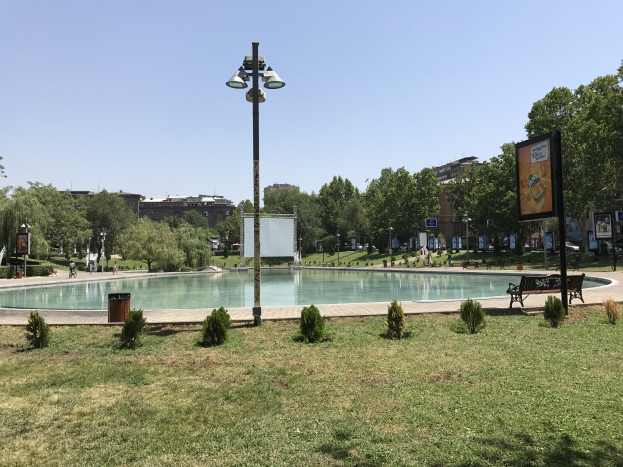 Großer Swimmingpool in einem Park umgeben von Grün, Strukturen und Menschen unter einem sichtbaren Himmel.