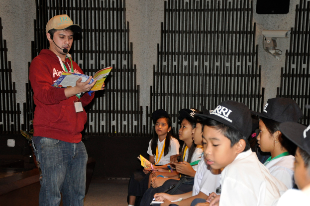 Ein Mann in einer marineblauen Kapuzenjacke und Jeans hält ein Buch und spricht in ein Mikrofon, mit ein paar Kindern in weißen T-Shirts und schwarzen Mützen auf der rechten Seite und einer Überwachungskamera an der Wand im Hintergrund.
