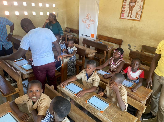 Kinder sitzen an Tischen und nutzen Tablets in einem Klassenzimmer mit Erwachsenen in der Nähe und Lehrplakaten an der Wand.