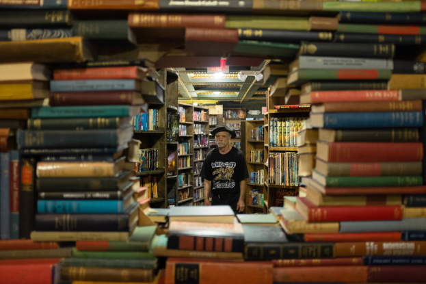 Ein Mann in einer Mütze, T-Shirt und Hose steht vor einem Bücherregal mit ordentlich angeordneten Büchern in einer Bibliothek.