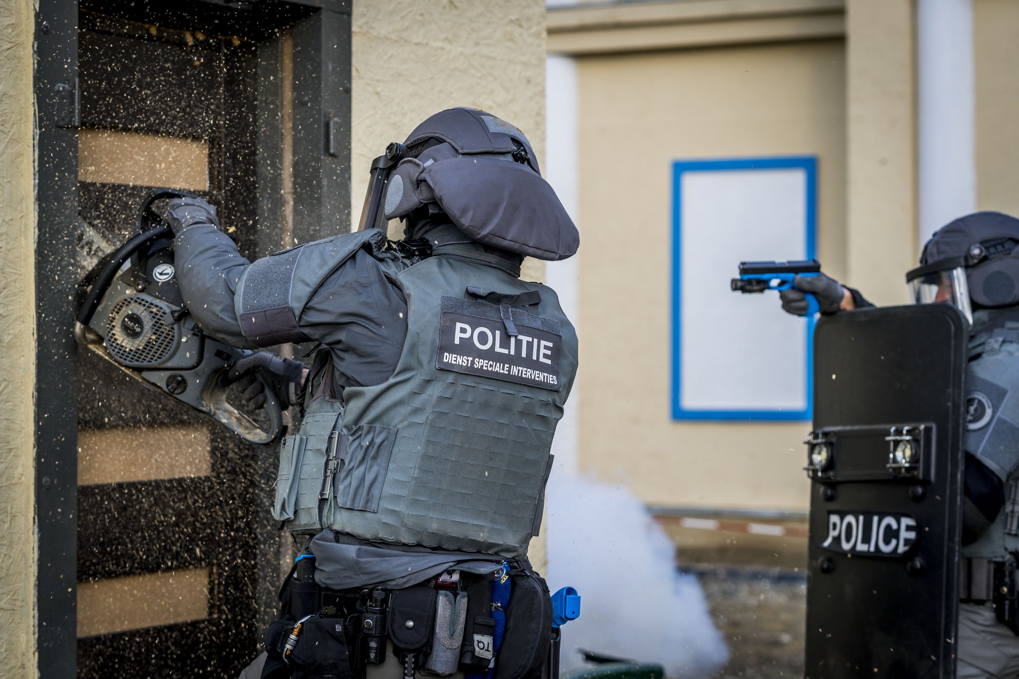 Zwei Polizisten in Einsatzkleidung stehen vor einem Gebäude, einer hält eine Waffe und der andere ein Objekt, mit einer Tür links und einer Tafel an der Wand im Hintergrund.