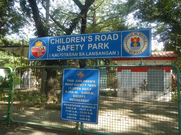 Metallzaun um einen Kinderverkehrssicherheitspark mit zwei blauen Informationsschildern, einem kleinen Dachhaus, Topfpflanzen, einer Baumgruppe und einem bewölktem Himmel.