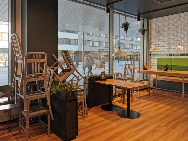 Ein Restaurant-Innenraum mit hölzernen Tischen und Stühlen in der Nähe eines großen Fensters, das eine städtische Berliner Straßenszene mit Fußgängern, Gebäuden und urbanen Elementen zeigt.