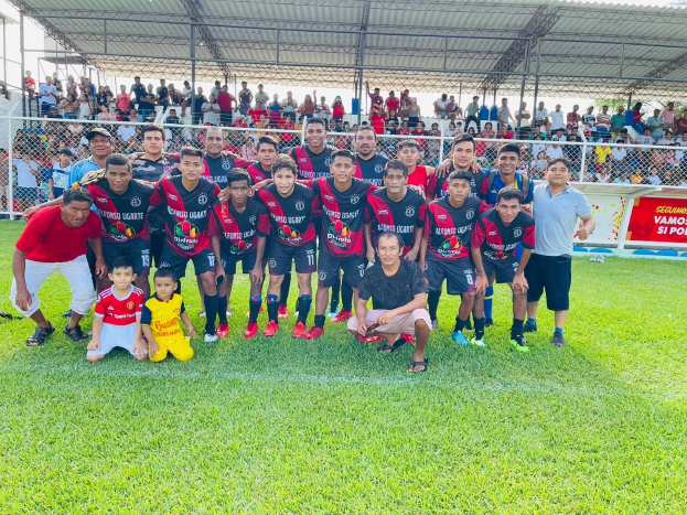 Gruppe von Männern, die auf einem Fußballfeld mit einem Banner feiern, das "Fotos Ugarte U-17 vs U-18" und Metallzäune unter einem bewölkten Himmel zeigt.