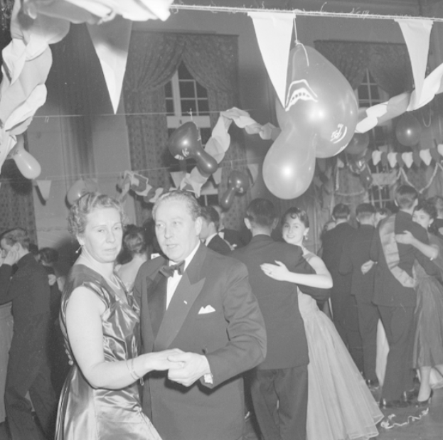 Ein Mann in einem schwarzen Blazer, weißem Hemd und schwarzen Hosen tanzt mit einer Frau auf einer Party, umgeben von Menschen, Ballons, Vorhängen und einem Fenster, alles in Schwarz-Weiß dargestellt.