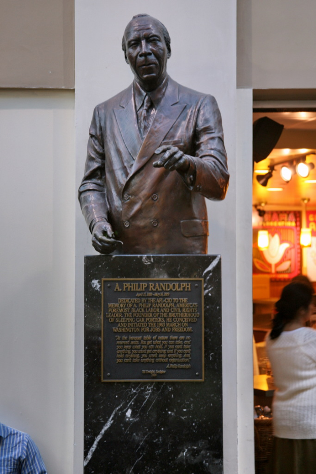 Eine Statue mit Text darunter, eine Frau rechts mit einigen Gegenständen und eine Person unten links, alles vor einer Wand.