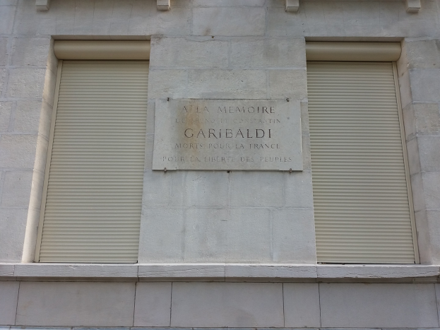 Ein Gebäude mit zwei Fenstern und geschlossenen Fensterläden sowie einer Gedenktafel an der Seite.