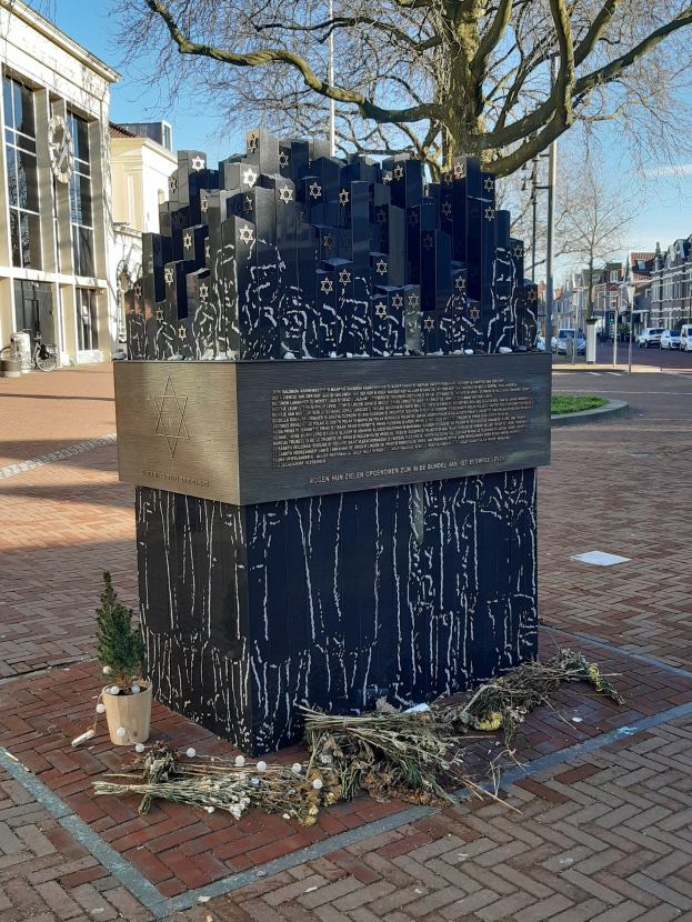 Denkmal in einem Stadtplatz, das Holocaust-Opfern gewidmet ist, mit einem Baum davor, Topfpflanze, verstreuten Blättern, Gebäuden, Straßenverkehr, Versorgungsmästen und Bäumen im Hintergrund.
