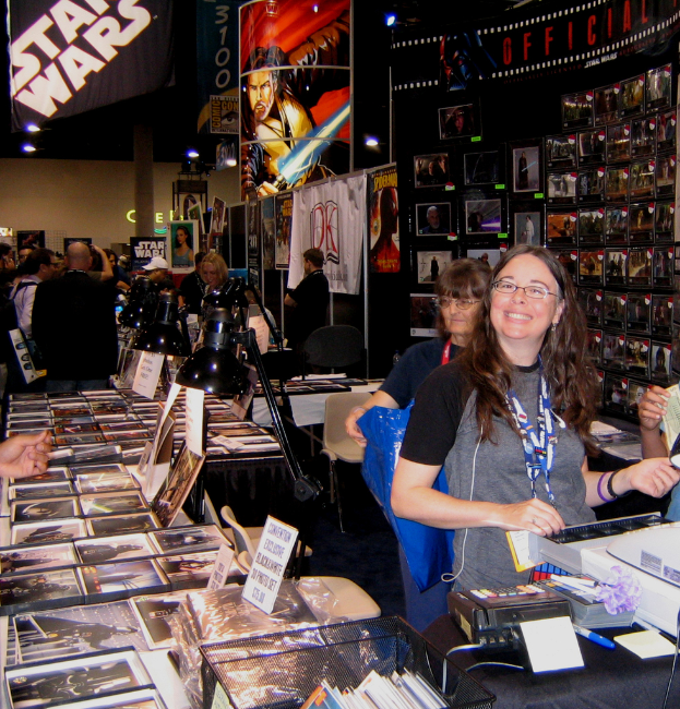 Eine Frau steht an der Kasse in einem belebten Geschäft, das mit Bildern, Büchern und präsentierten Plakaten gefüllt ist, im Hintergrund kaufen Kunden ein.