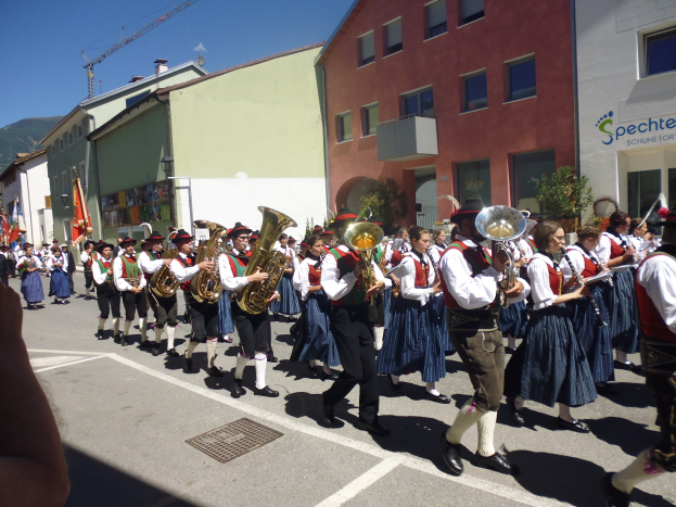 Eine Gruppe von Menschen in traditioneller bayrischer Tracht, die auf der Straße musizieren, während sie durch eine Straße mit Gebäuden marschieren, einige halten Fahnen, mit einem Hügel und einem klaren blauen Himmel im Hintergrund.