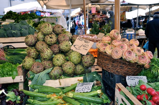 Mit Obst, Gemüse und kleinen Schildern gefüllte Körbe vor Zelten und ein paar Menschen im Hintergrund.