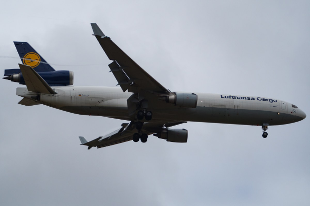 Lufthansa Cargo Boeing 777-300ER im Flug mit sichtbarem Text auf dem Rumpf, ausgefahrenen Tragflächen, aktiven Triebwerken vor einem Hintergrund aus weißen Wolken.