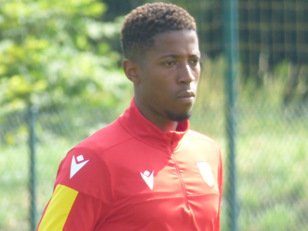 Ein junger Mann in einem roten und gelben T-Shirt steht auf einem Fußballfeld, umgeben von einem Zaun und Bäumen, und sieht entschlossen während eines Trainings.