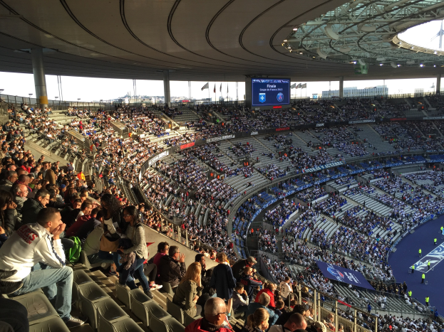 Großer Menschenauflauf in einem Stadion bei einem Fußballspiel, mit einer Bühne rechts, Fahnen, Stangen, einem Bildschirm und der Allianz Arena in München, Deutschland im Hintergrund.