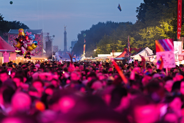 Eine große Menschenmenge auf einem nächtlichen Musikfestival, beleuchtet von bunten Lichtern, mit Zelten, Fahnen und Pfählen im Vordergrund, Bäumen und einem Turm im Hintergrund und einem klaren Nachthimmel.