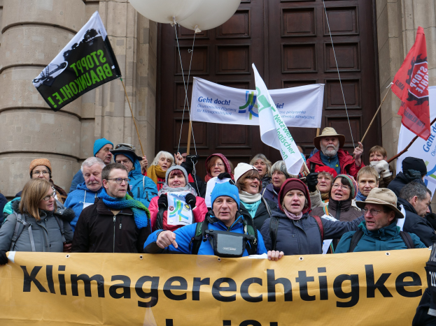 Eine Gruppe von Menschen nimmt an einem Klimastreik in Deutschland teil, hält ein Banner, Flaggen und Luftballons vor einem Gebäude mit Säulen und einer Tür.