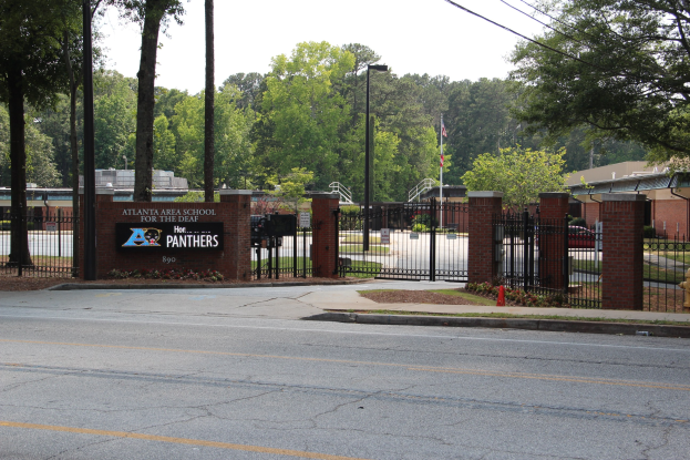 Einfahrt zu einer Schule mit einem Schild, umgeben von Bäumen, Straßenlaternen, Fahrzeugen und Gebäuden unter einem klaren blauen Himmel.