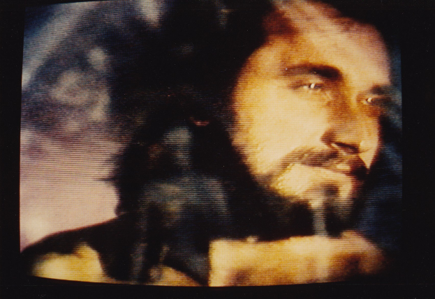 Ein Mann mit langem dunklem Bart und Haar, der ein weißes Hemd trägt, erscheint auf einem Fernsehbildschirm mit einem ernsten Gesichtsausdruck, seine Augen und geöffneten Lippen sind auf den Betrachter gerichtet, vor einem dunklen Hintergrund.