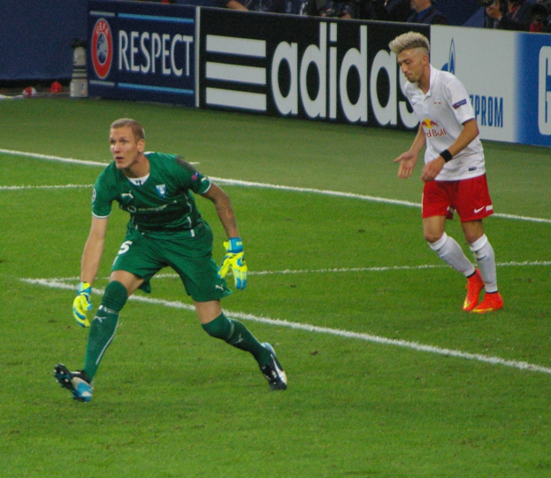 Zwei M├Ąnner spielen Fussball auf einem gr├╝nen Feld, umgeben von einer Menschenmenge, einige machen Fotos, mit Texttafeln im Hintergrund.