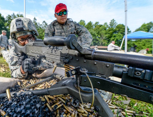 Eine Person auf einem Armee-Trainingsgelände schießt mit einem Gewehr, während eine andere Person mit einer roten Mütze daneben steht, während im Hintergrund Bäume, Zelte und Menschen zu sehen sind, unter einem klaren, sonnigen Himmel, mit Patronenhülsen und Gras im Vordergrund.