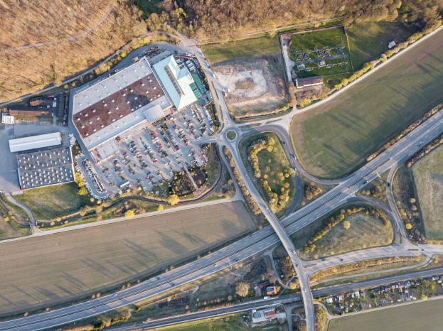 Luftaufnahme eines großen Gebäudes auf einem Feld, umgeben von Straßen, Fahrzeugen, Bäumen und Gras, mit einem Logo, das es als deutsche Automobilindustrie-Zentrale identifiziert.