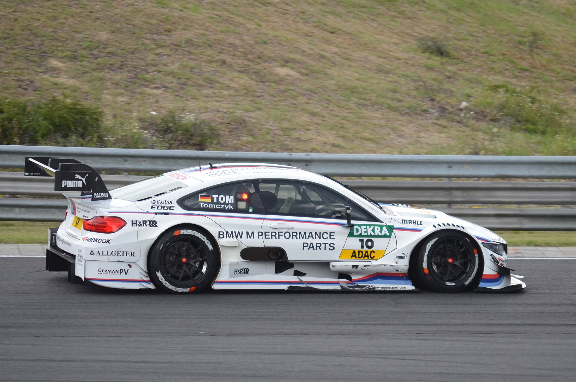 BMW M4 DTM-Rennwagen auf einer Rennstrecke mit grünem Gras und einer Absperrung im Hintergrund.