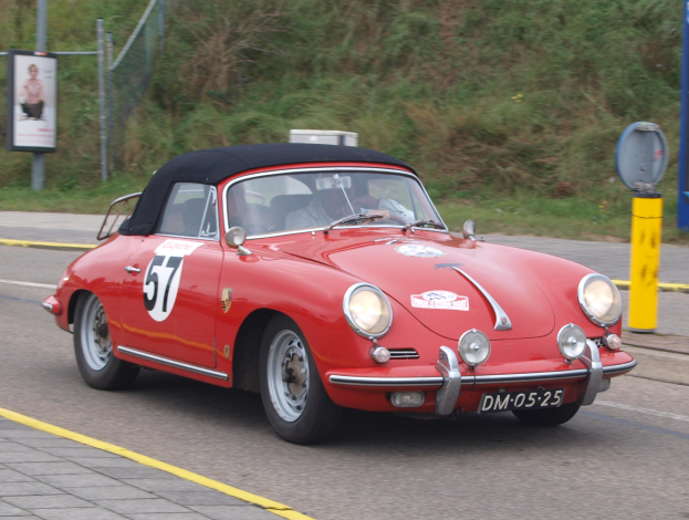 Roter Porsche 356 Speedster mit zwei Insassen auf einer Rennstrecke, Pfahl mit angebrachten Schild auf der rechten Seite, Pflanzen und Zaun im Hintergrund.